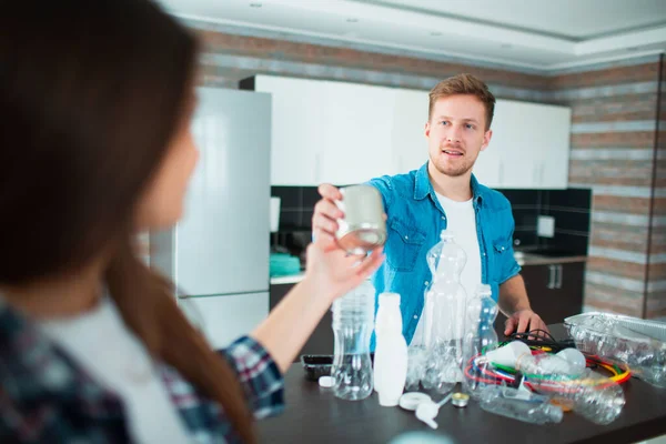 Una familia joven clasifica los materiales en la cocina para su reciclaje. Los materiales reciclables deben separarse. La esposa le da a su marido una lata vieja para comestibles. Arreglan toda la basura de la casa. — Foto de Stock