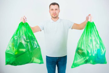 Genç adam beyaz arka planda izole edilmiş. Adam içinde çöp ve atık olan iki plastik torba tutuyor. Yeniden dönüşüm ve yeniden kullanım için hazırlanılıyor.