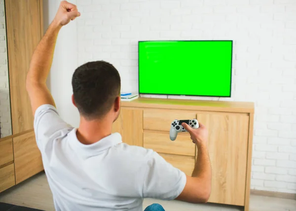 Giovane uomo seduto nella stanza durante la quarantena. Ragazzo che gioca ai giochi per computer usando joystick e tv. Schermo verde. Giocare da solo e fare il tifo. Vista posteriore . — Foto Stock