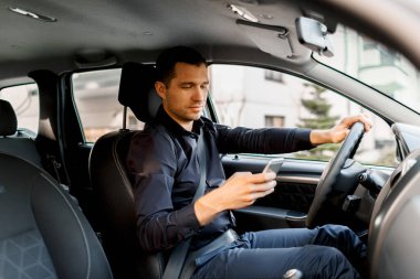 Kendi arabasını kullanan koyu renk gömlekli genç bir adam akıllı telefon ya da telefon kullanıyor. Çoklu görev kavramı