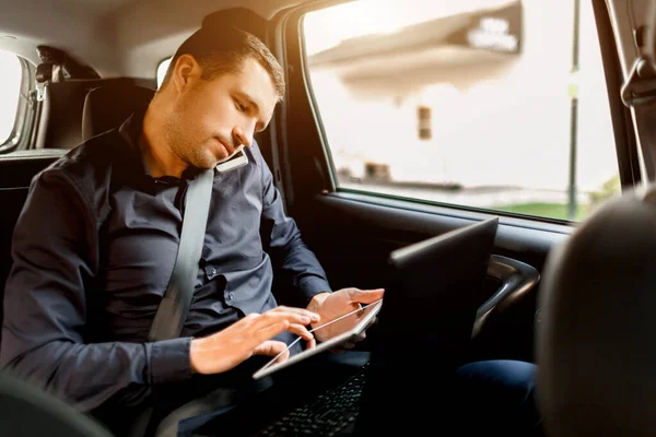 Un uomo d'affari impegnato in un taxi. Concetto multitasking. I passeggeri viaggiano sul sedile posteriore e lavorano simultaneamente. Parla su uno smartphone e utilizza un computer portatile e tablet . — Foto Stock