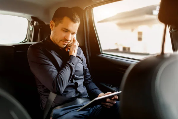 Un uomo d'affari impegnato in un taxi. Concetto multitasking. I passeggeri viaggiano sul sedile posteriore e lavorano simultaneamente. Parla su smartphone e utilizza tablet pc . — Foto Stock