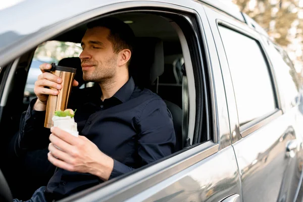 Останавливаюсь перекусить. Человек ест закуски в машине и пьет кофе или чай . — стоковое фото
