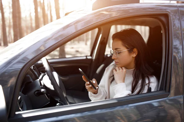 A jovem está sentada num carro. Modelo morena bonita está usando um smartphone ou telefone — Fotografia de Stock