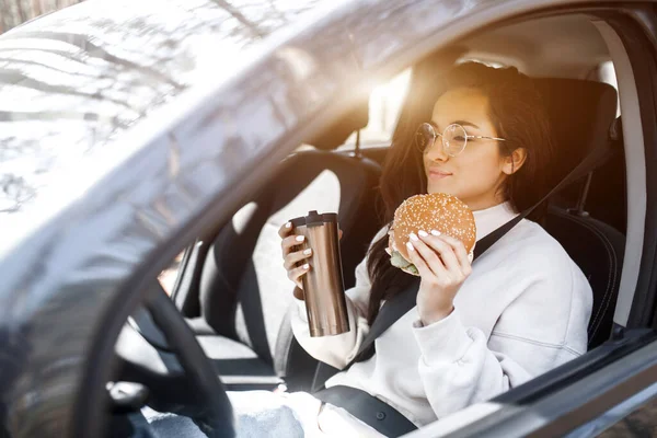 Bella modella bruna pranza in macchina. Una donna mangia un hamburger e beve caffè o tè da un thermos. Il cibo sulla strada . — Foto Stock