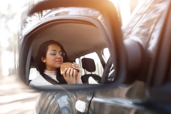 Modelo morena bonita tem almoço em um carro. Mulher come um hambúrguer e bebe café ou chá de uma garrafa térmica. Comida no conceito de estrada . — Fotografia de Stock