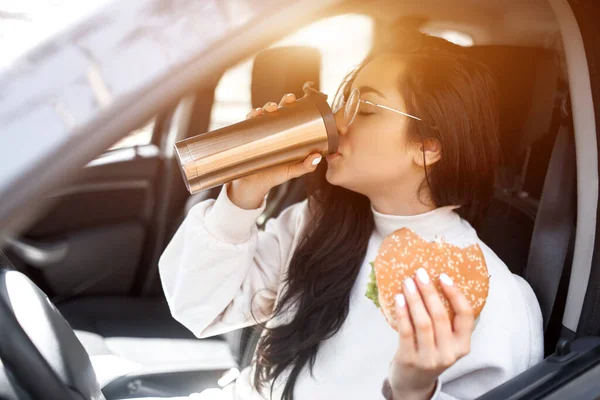 Schöne brünette Modell hat Mittagessen in einem Auto. Frau isst Hamburger und trinkt Kaffee oder Tee aus der Thermoskanne. Essen auf der Straße. — Stockfoto