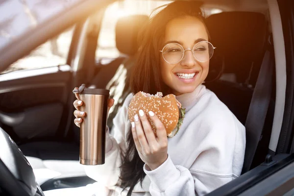 Modelo morena bonita tem almoço em um carro. Mulher come um hambúrguer e bebe café ou chá de uma garrafa térmica. Comida no conceito de estrada . — Fotografia de Stock