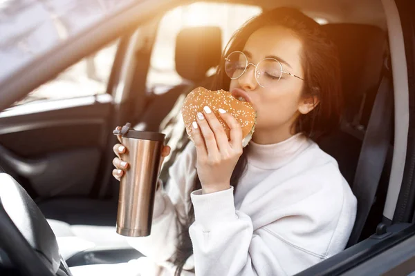 Mat på vägen konceptet. Kvinnan äter en hamburgare och dricker kaffe eller te från en termos. Livsmedel på väg konceptet. — Stockfoto
