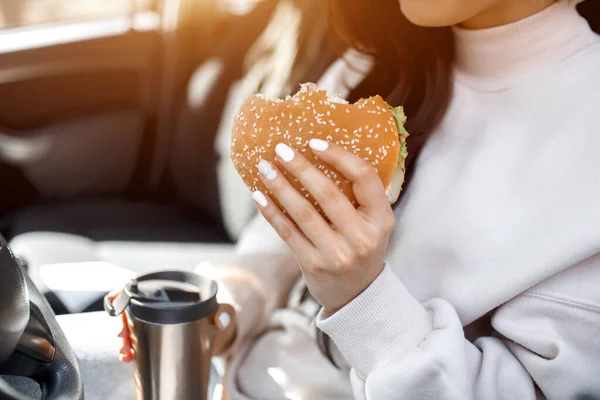 Mat på vägen konceptet. Kvinnan äter en hamburgare och dricker kaffe eller te från en termos. Livsmedel på väg konceptet. — Stockfoto