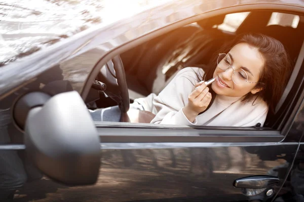 Een jonge vrouw in een auto. Ze schildert haar lippen met lippenstift in de achteruitkijkspiegel — Stockfoto