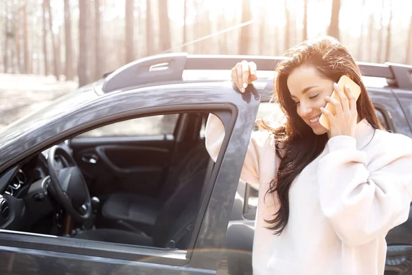 Uma bela jovem de cabelos pretos está de pé perto de um carro em uma floresta ou parque. As modelos femininas têm uma viagem para fora da cidade em um cruzamento. Ela fala no telefone ou smartphone e mantém uma garrafa térmica em seu — Fotografia de Stock