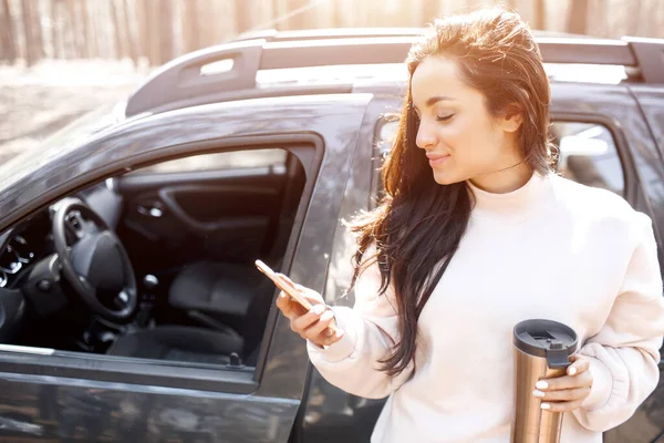 Uma bela jovem de cabelos pretos está de pé perto de um carro em uma floresta ou parque. As modelos femininas têm uma viagem para fora da cidade em um cruzamento. Ela fala no telefone ou smartphone e mantém uma garrafa térmica em seu — Fotografia de Stock