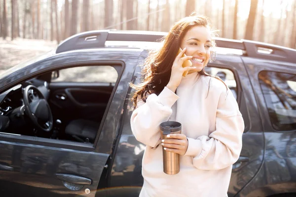 一位漂亮的黑头发年轻女子站在森林或公园里的一辆汽车旁边。女模特儿乘越野车出城旅行.她在电话或智能手机上说话，手里拿着一个热水瓶 — 图库照片