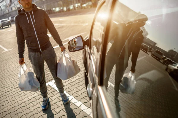 Egy férfi áll a parkolóban egy bevásárlóközpont vagy egy bevásárlóközpont közelében. Mindent sikeresen megvett a szupermarketben. Élelmiszerekkel, élelmiszerzöldségekkel és gyümölcsökkel, tejtermékekkel töltött műanyag zacskók. — Stock Fotó