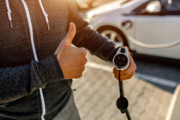 Homem segurando cabo de carregamento de energia para carro elétrico no parque de estacionamento ao ar livre. O motorista um polegar para cima — Fotografia de Stock