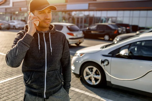 Un uomo aspetta mentre la sua auto elettrica si carica a pagamento in un parcheggio vicino a un centro commerciale . — Foto Stock