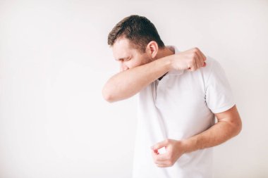 Genç hasta bir adam beyaz arka planda izole edilmiş. Yüksek sesle öksür ve ağzını dirsekle kapat. Acı verici öksürük. Stüdyodaki adam tek başına. Hastalıktan acı çek.