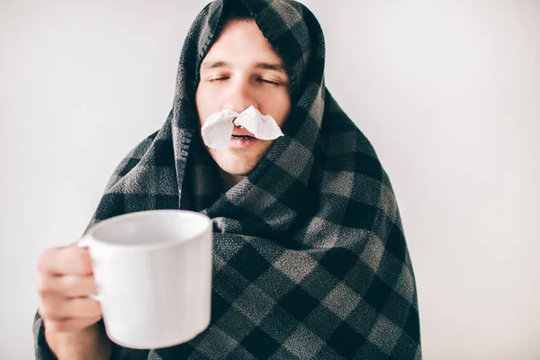 Young sick man isolated over white background. Guy covered with blanket. Holding cup of tea with closec nose tissues. Drink medicine for better self condition. Sick and ill person with disease.