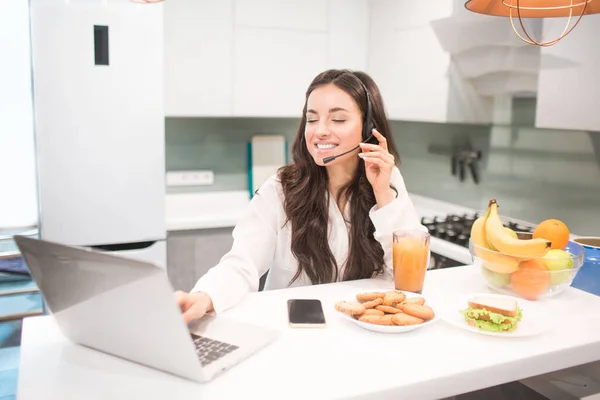 Beautiful black-haired woman works from home and uses headphones with a headset. An employee sits in the kitchen and has a lot of work on a laptop and tablet and has video conferencing and meetings.