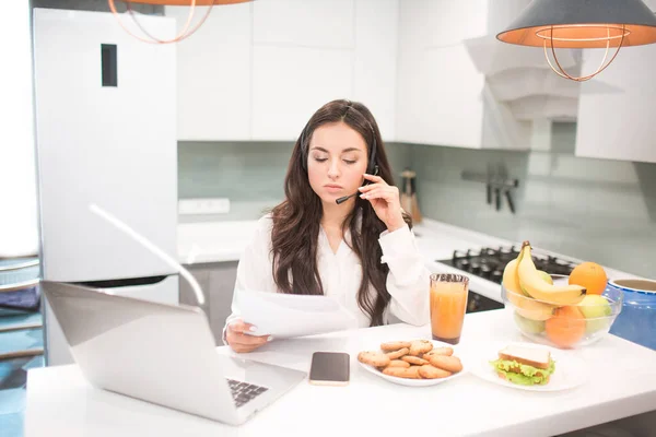 Piękna czarnowłosa kobieta pracuje w domu i używa słuchawek ze słuchawkami. Pracownik siedzi w kuchni i ma dużo pracy na laptopie i tablecie i ma wideokonferencje i spotkania. — Zdjęcie stockowe