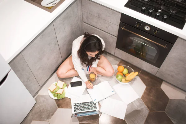 Bela mulher de cabelos negros trabalha em casa. Um funcionário se senta na cozinha e tem muito trabalho em um laptop e tablet e tem videoconferência e reuniões — Fotografia de Stock