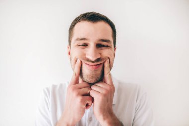 Genç adam beyaz arka planda izole edilmiş. Zorla gülümseyen bir adamın yüz resmi. Dudakların kenarlarında parmak tutmak.