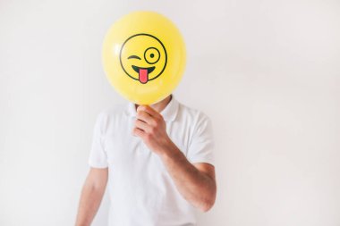 Genç adam beyaz arka planda izole edilmiş. Gömlekli adam ön yüzünde sarı balon tutuyor. Dili dışarıda boyanmış emoji. Yüzünü gizle ya da ört.
