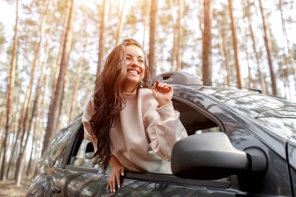 Gelukkige jonge vrouw klom uit een autoraam. Reizen met de auto in het bos. Een reisje. Vakantie met de auto — Stockfoto