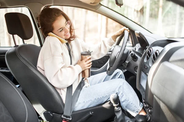 A jovem está sentada num carro. Modelo morena bonita está usando um smartphone ou telefone — Fotografia de Stock