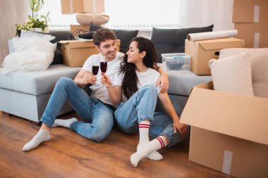 Genç mutlu çift şarap içiyor, yeni eve taşınmayı kutluyor ve kutuların arasında oturuyor..