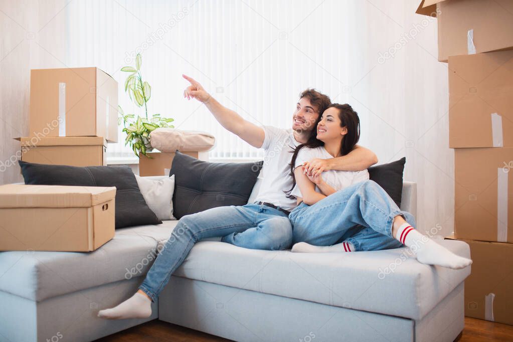 a happy young married couple moves to new apartment. They are sitting on the sofa and looking at their new home.