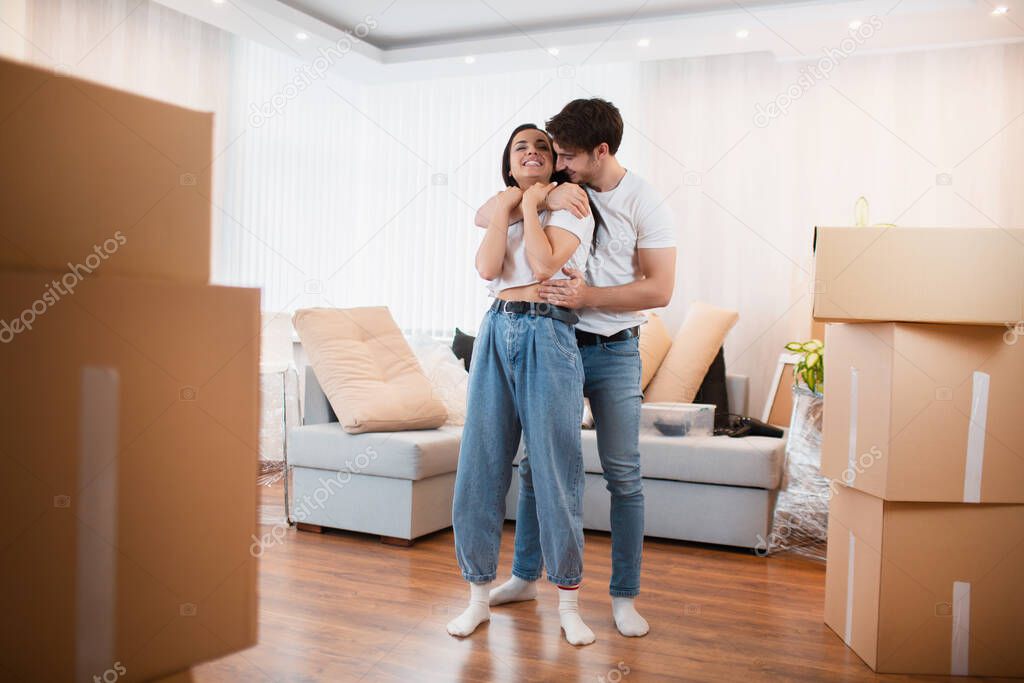 A young married couple in the living room in the house stand near unpacked boxes. hey are happy about new home. Moving, buying a house, apartment concept.
