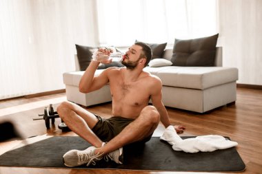 Genç adam karantina sırasında spor yapıyor. Spor figürlü tişörtsüz adam bacak bacak üstüne atıp şişeden su içiyor. Antrenmandan sonra dinlen..