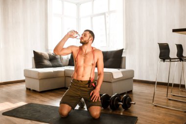 Genç adam karantina sırasında spor yapıyor. Egzersizden sonra dinlenin. Tişörtsüz sporcu adam mindere oturup plastik şişeden su içiyor. wokrout sonrası duraklat.