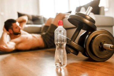 Genç adam evde kanepede karın kası egzersizi yapıyor. Dairesinde spor yapan, sıkı çalışan tişörtsüz bir sporcunun görüntüsünü kes. Dambıllar yerde yatıyordu. İyileşmeye çalışıyorum.