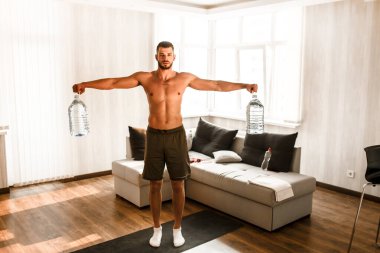 Genç adam karantina sırasında spor yapıyor. Spor figürleri olan tişörtsüz bir sporcu doğaçlama pullu bir sporcu. Dambıl yerine büyük plastik su şişeleri kullanıyor.