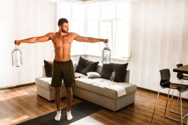 Genç adam karantina sırasında spor yapıyor. Spor figürleri olan tişörtsüz bir sporcu doğaçlama pullu bir sporcu. Dambıl yerine büyük plastik su şişeleri kullanıyor.