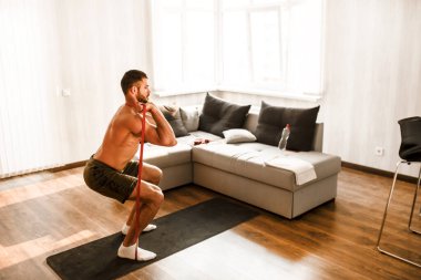 Tişörtsüz sporcu eve spor olsun diye gidiyor. Genç adam karantina sırasında spor yapıyor. Yoga minderinin üzerinde dur ve direnç bandını kullanarak bacaklarını arkaya uzat..