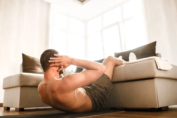 Joven haciendo ejercicios abdominales en casa en el sofá. Vista cortada del hombre sin camiseta deportista en actividad de entrenamiento en su apartamento. pesas en las fotos que yacen en el suelo. Tratando de mejorar . — Foto de Stock