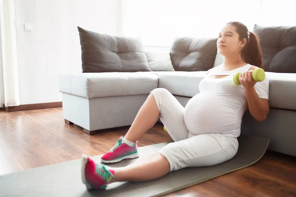 Elinde halterlerle yoga minderinde oturan hamile bir kadın. Hamile kadınlar için spor ve spor. Yakında doğum — Stok fotoğraf