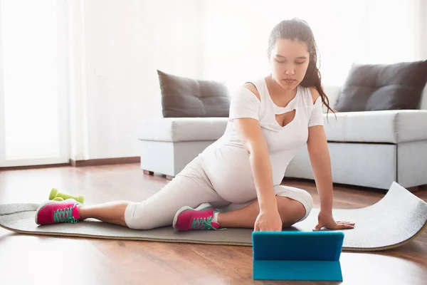 Gravid kvinna sitter på en yogamatta och använder en mobil applikation på en tablett för sport under graviditeten — Stockfoto