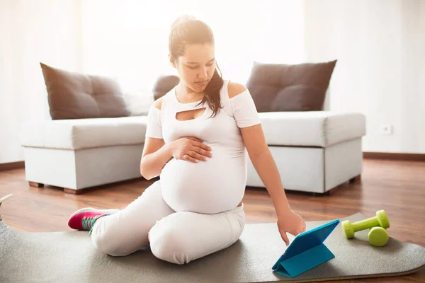 Hamile kadın yoga minderinde oturur ve hamilelik sırasında spor için mobil bir uygulama kullanır. — Stok fotoğraf