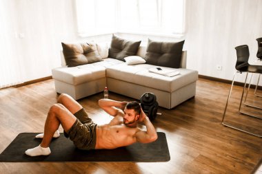 Genç adam spor için evine git. Tişörtsüz bir sporcunun yoga minderinde karın kası egzersizi yaparken fotoğrafı. Birinci sınıfların vücut ısıtması. Güneşli bir dairede tek başına geriniyorsun.