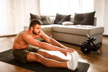Genç adam spor için evine git. Spor figürlü tişörtsüz bir adam. Çekiciyle bacaklarına kadar esniyor. Boş bir dairede tek başına alıştırma yapıyorsun. Formda kalmaya ve sağlıklı kalmaya çalış..