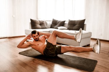 Genç adam spor için evine git. Tişörtsüz bir sporcunun yoga minderinde karın kası egzersizi yaparken fotoğrafı. Birinci sınıfların vücut ısıtması. Güneşli bir dairede tek başına geriniyorsun.