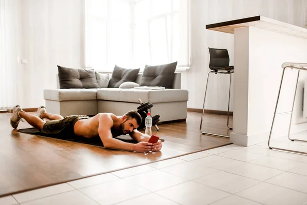 Ung vanlig man går in för sport hemma. T-shirtless kille idrottsman har vila på golvet och använda smartphone. Freshman i träning avkopplande efter träning. Ensam i lägenheten — Stockfoto