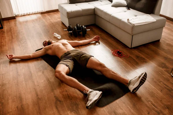 El tipo está entrenando en casa y muy cansado después del entrenamiento. Está tumbado en una esterilla de yoga sudoroso y sin camiseta — Foto de Stock