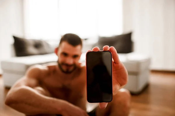 ハードウォーキングTシャツのない男スポーツマンは、スマートフォンのクローズアップを示しています。ソファの上で家で運動してる若い男. — ストック写真