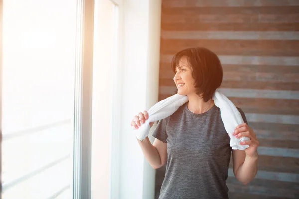 Adulte en forme femme mince a séance d'entraînement à la maison. Une femme âgée forte et bien construite se tient seule dans la pièce après l'exercice. Serviette blanche autour du cou. Repose-toi et relaxe . — Photo